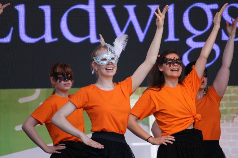 Vier Mädchen in orange Shirts und schwarzen Röcken mit Masken auf der Lindenfest-Bühne, zwei heben die Arme.