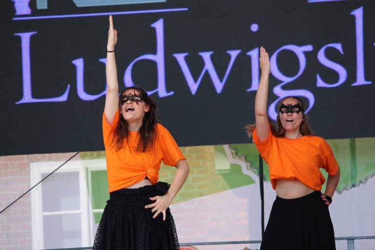 Zwei junge Darstellerinnen in orangen Shirts und schwarzen Röcken mit Masken beim Lindenfest Ludwigslust Auftritt.