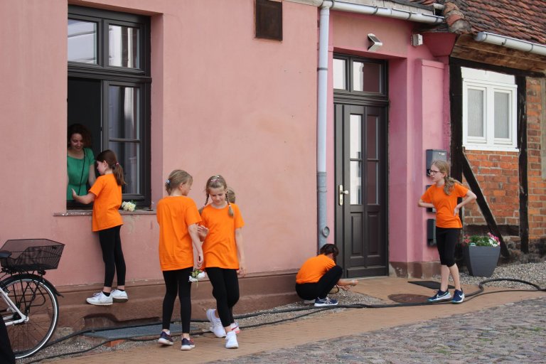 Gruppe von Kindern in orangefarbenen Musicalschul-T-Shirts bei Außenaktivität vor rosa Gebäude beim Lindenfest Ludwigslust.