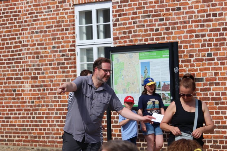 Mann führt Gruppe von Kindern bei Stadtführung am Lindenfest Ludwigslust vor Informationstafel an Backsteingebäude.