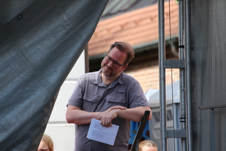 Mann mit Brille in grauem Hemd hält Papier auf Bühne beim Lindenfest Ludwigslust der Musicalschule Ahrensburg.
