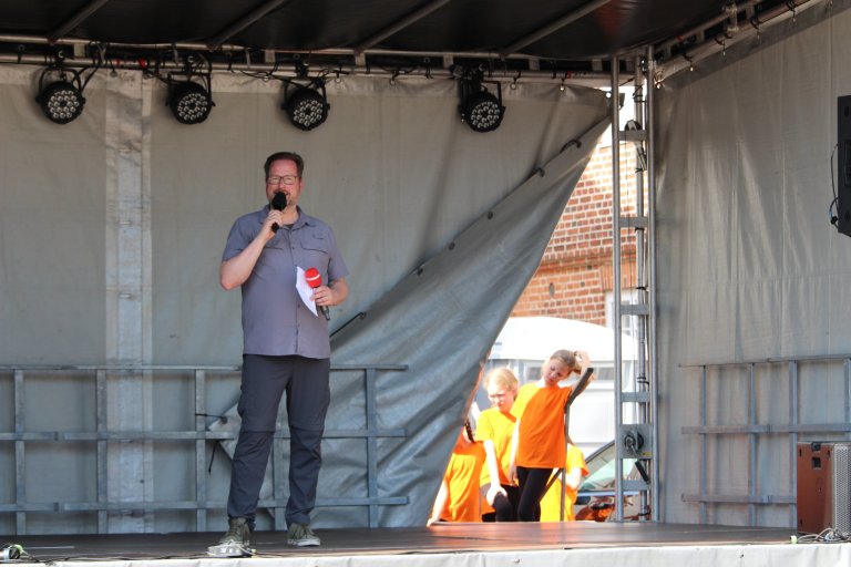 Mann mit Mikrofon auf Bühne, Kinder in orangenen Westen im Hintergrund beim Lindenfest Ludwigslust.