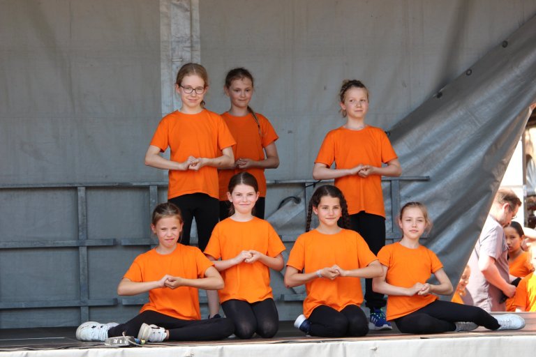 Kindertheatergruppe in orangefarbenen Shirts beim Lindenfest Ludwigslust sitzt und steht auf einer Bühne.