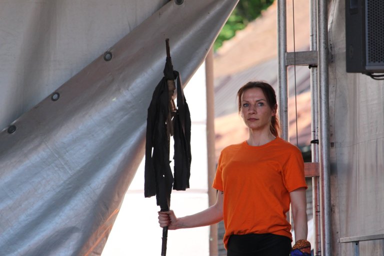 Junge Schauspielerin in orangem Shirt hält schwarze Theaterfahne beim Lindenfest Ludwigslust der Musicalschule Ahrensburg.