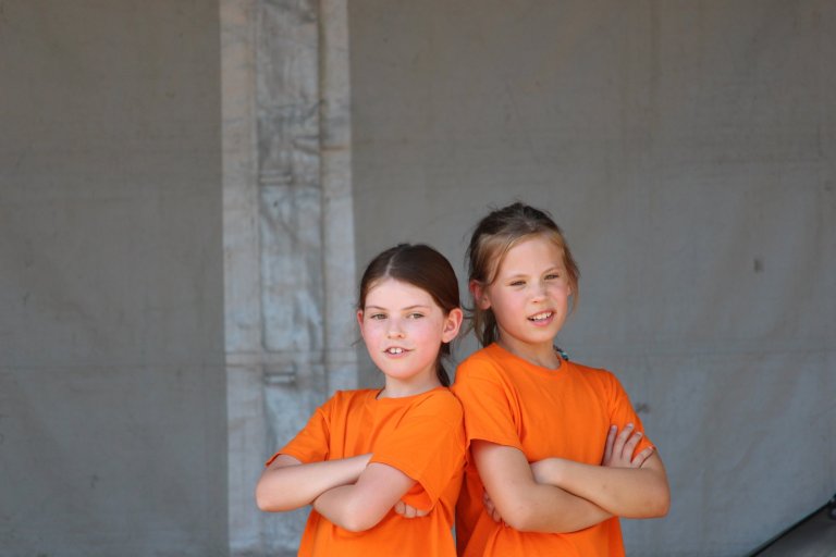 Zwei lächelnde Mädchen in orangefarbenen T-Shirts stehen mit verschränkten Armen vor grauer Wand.