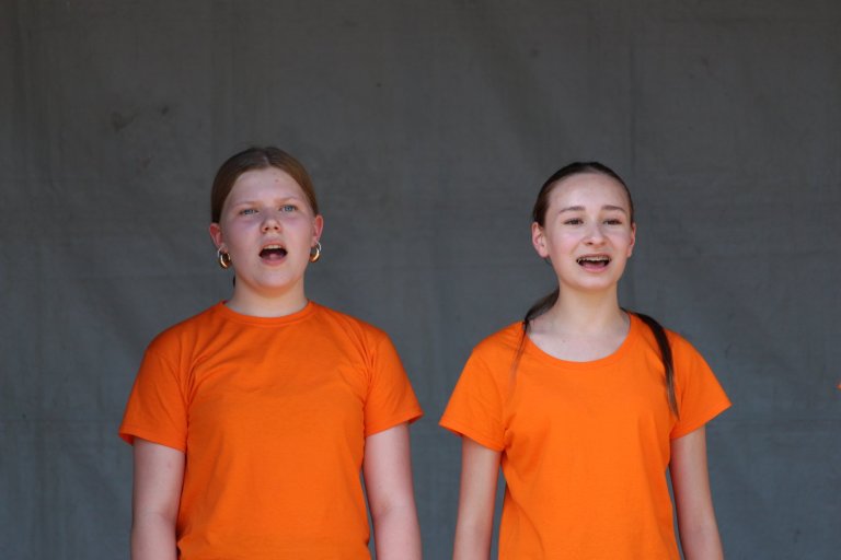 Zwei Mädchen in orangenen T-Shirts singen gemeinsam vor grauem Hintergrund beim Lindenfest Ludwigslust.