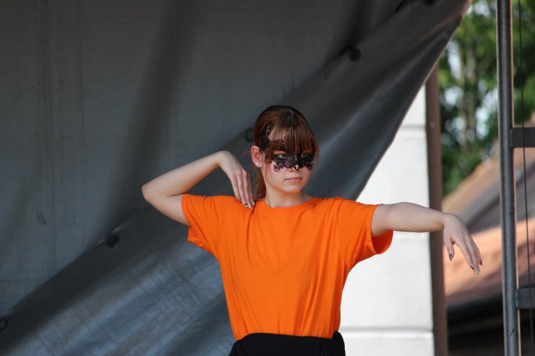 Junge Performerin in orangem Shirt mit schwarzer Spitzenmaske beim Lindenfest Ludwigslust Auftritt der Musicalschule Ahrensburg.
