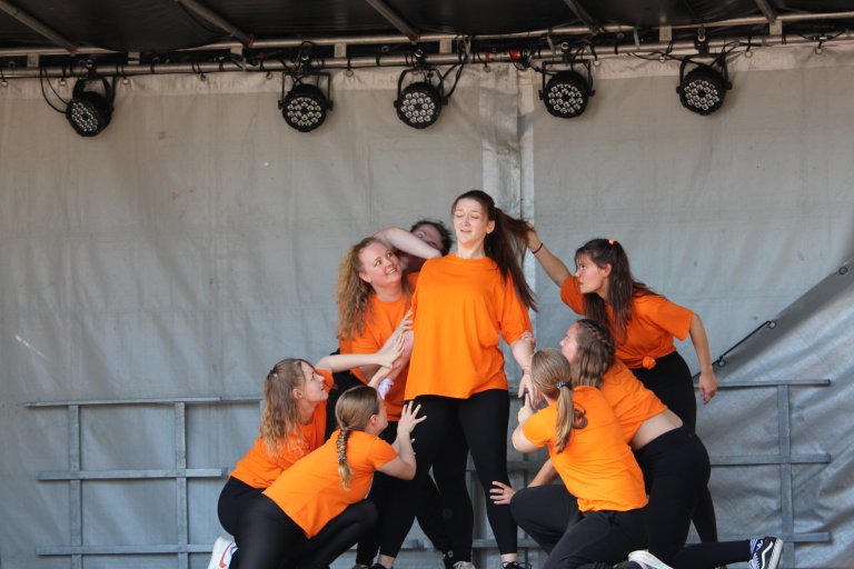 Gruppe junger Tänzerinnen in orangefarbenen Shirts auf einer Bühne mit professioneller Bühnentechnik.