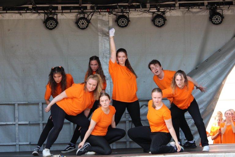 Tanzgruppe in orangefarbenen Shirts präsentiert synchrone Choreografie auf Bühnenbühne mit Scheinwerfern.