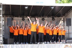 Musicalschüler in orangen und gelben Shirts performend auf Bühne mit Armen hochgestreckt, synchrone Tanzchoreographie.