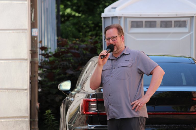 Mann mit Brille hält Mikrofon vor Auto in Garagensetting, trägt lila Hemd.